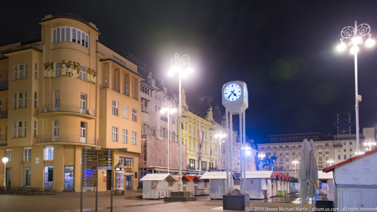Ban Jelačić Square at night with the tall blue-faced public clock, bright streetlamps, closed stalls, and historic buildings.