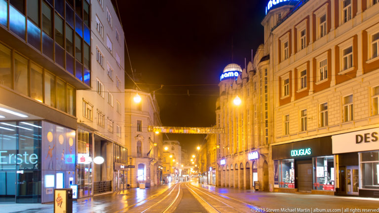 Straight tram-lined street in central Zagreb at night, with glowing shop signs, overhead wires, and historic commercial buildings.