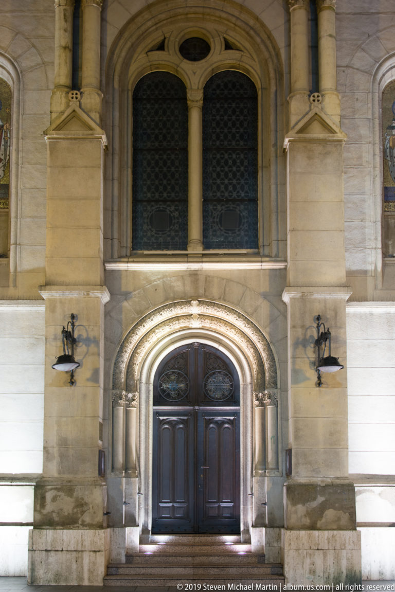 Illuminated church entrance with carved round arch, dark double doors, paired upper windows, and symmetrical wall lamps.