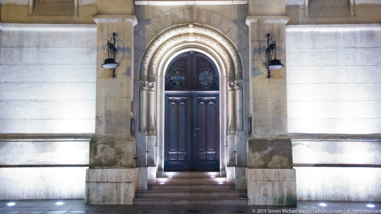 Dark wooden church door within a carved round-arched stone portal, flanked by wall lamps and lit from below.