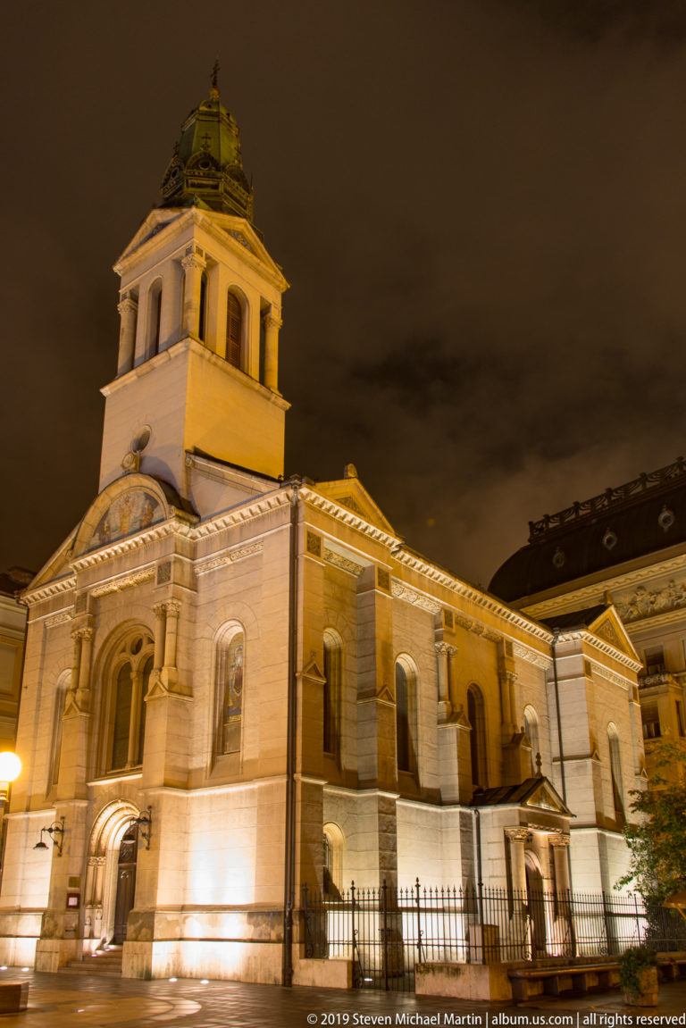 Oblique nighttime view of the illuminated Serbian Orthodox Cathedral of the Transfiguration behind an iron fence.