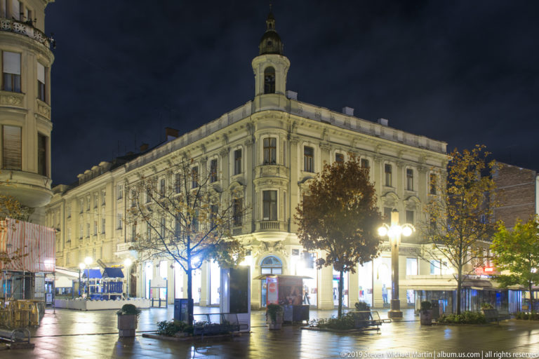 Illuminated multi-story corner building with rooftop cupola beside a quiet square, trees, kiosks, and wet pavement at night.