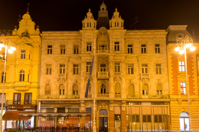 Ornate historicist building with balconies, arched windows, central pediment, and ground-floor cafés illuminated on Ban Jelačić Square.