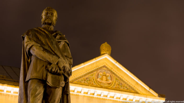 Low-angle rear view of the Petar Preradović statue against a brightly lit church pediment at night.