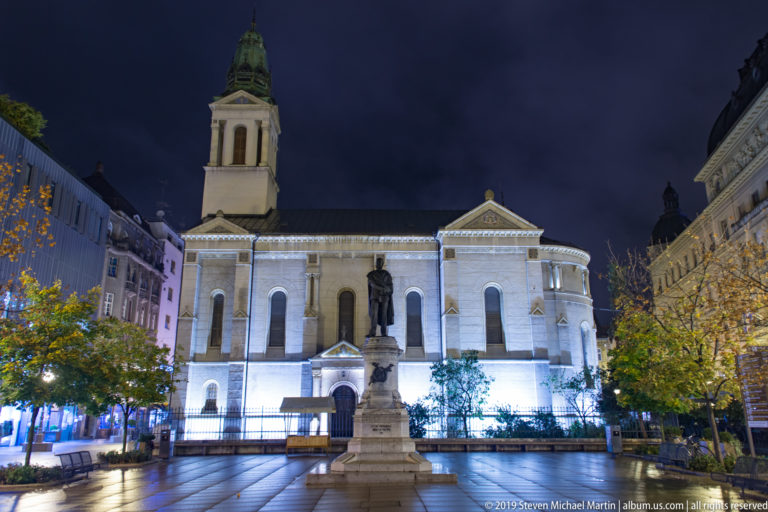 Illuminated Orthodox church and bell tower behind the Petar Preradović monument on a wet Zagreb square at night.