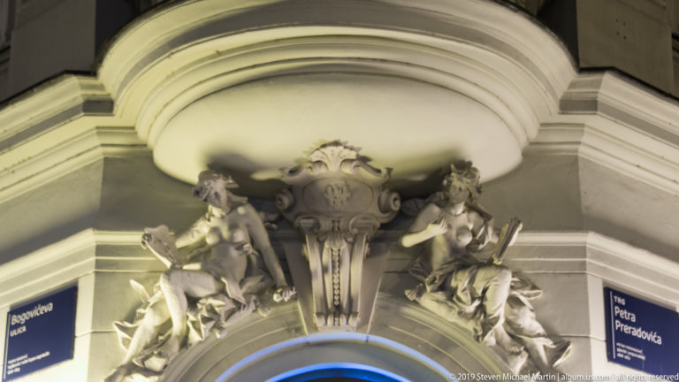 Sculpted figures and a central cartouche above an arched doorway, illuminated from below at night.