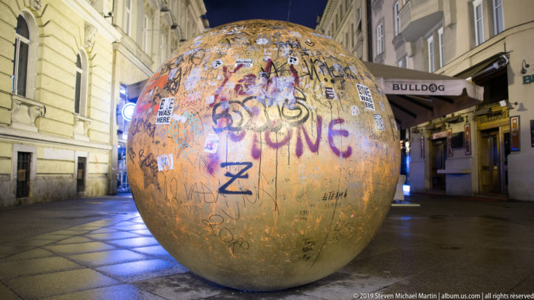 Graffiti-covered bronze sphere centered on a pedestrian street at night, flanked by illuminated shops and historic buildings.