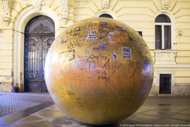 Large graffiti-covered bronze sphere on a paved Zagreb street at night, with an ornate pale building behind it.