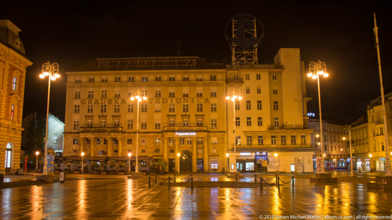 Gradska štedionica building across wet Ban Jelačić Square, with decorative streetlamps and reflections at night.