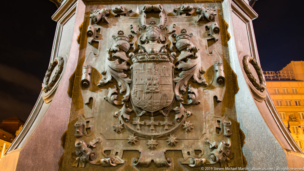 Large ornate metal coat of arms with shield, crest, scrollwork, and relief emblems mounted on polished stone at night.