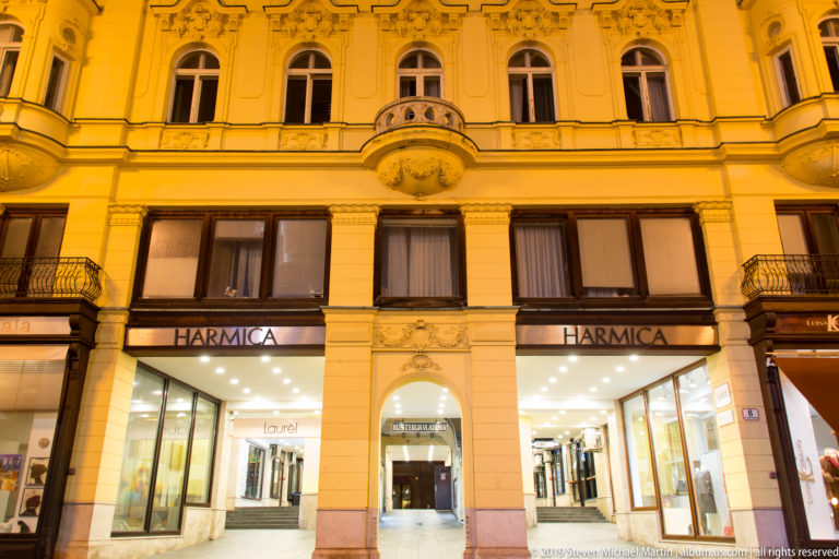 Harmica passage entrance with illuminated shops below an ornate historic façade, the word HARMICA visible on both sides.