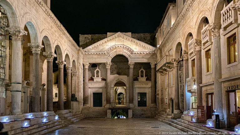 Frontal nighttime view of the Peristyle with illuminated colonnade, central arched façade, stone paving, and historic buildings.