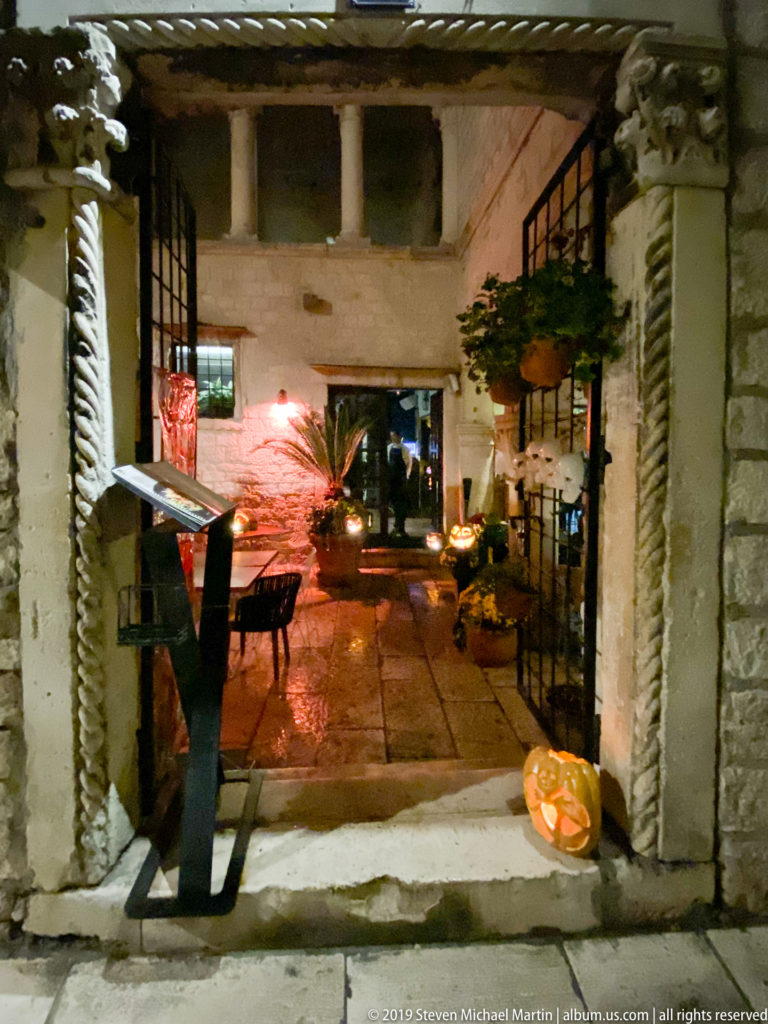 Small stone courtyard at night with café tables, chairs, potted plants, and warm lighting beneath old masonry walls.