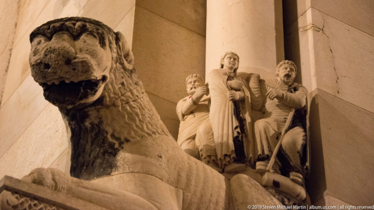 Weathered stone sphinx in the Peristyle foreground with illuminated carved figures and columns rising behind it at night.