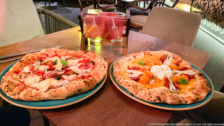 Two pizzas with varied toppings sit on ceramic plates at a small restaurant table with drinks and chairs behind them.