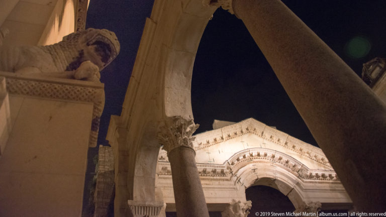 Peristyle columns and arch frame the illuminated façade of Saint Domnius Cathedral against a black night sky.