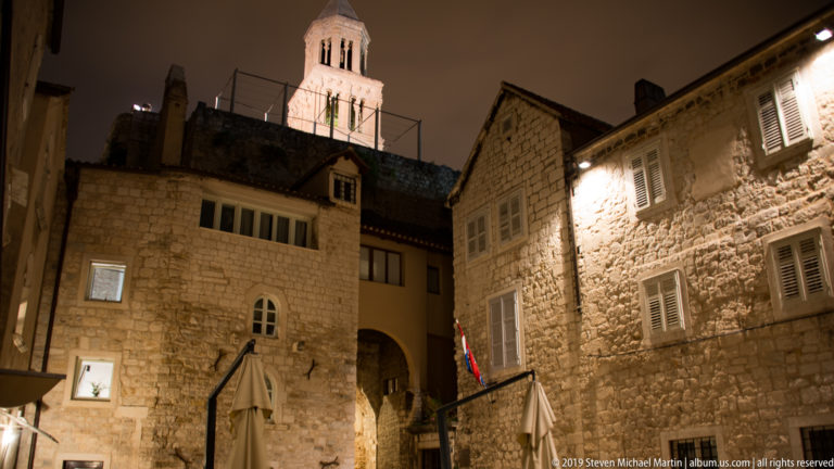Stone courtyard with shuttered windows and a small arch, with the illuminated Cathedral of Saint Domnius bell tower above the roofs.