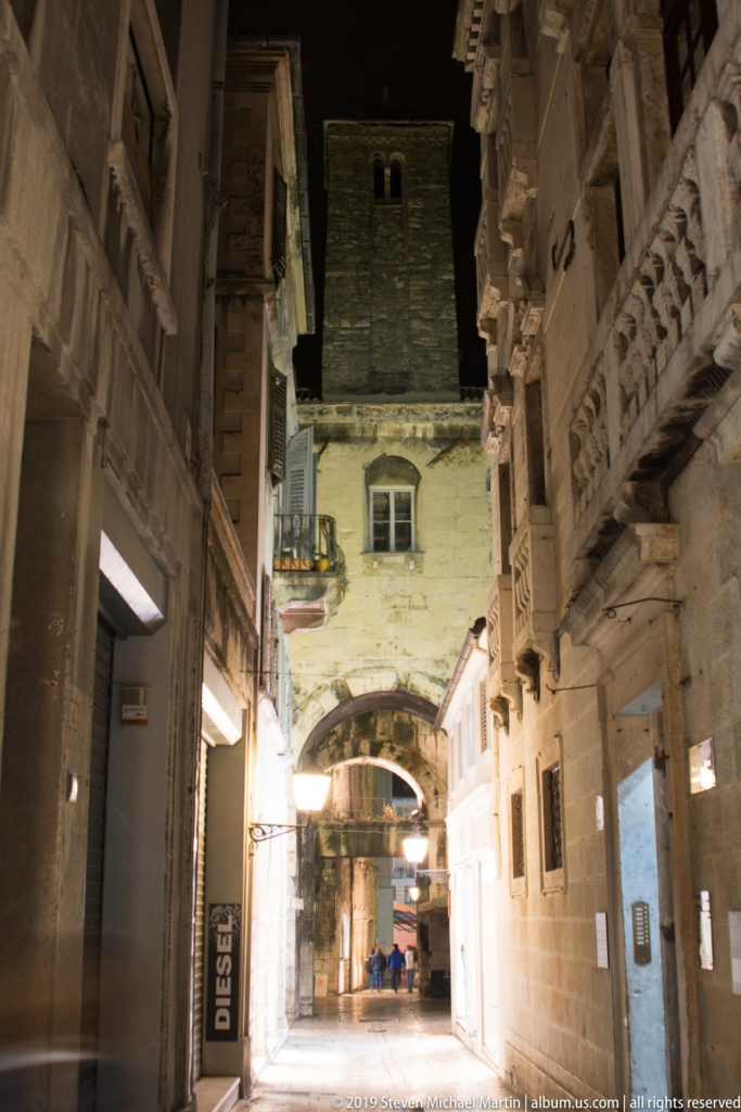 Illuminated stone passage with carved balcony details and an arch leading deeper into the Split City Museum complex.