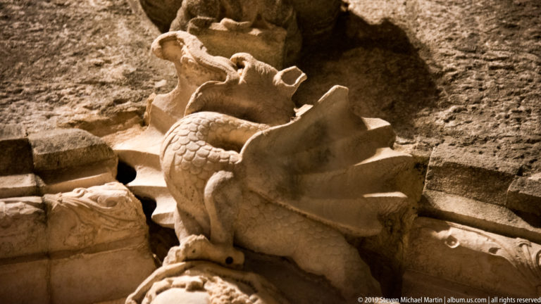 Weathered carved stone fantastic beast with scales, raised wings, open jaws, and broken surrounding fragments under warm light.