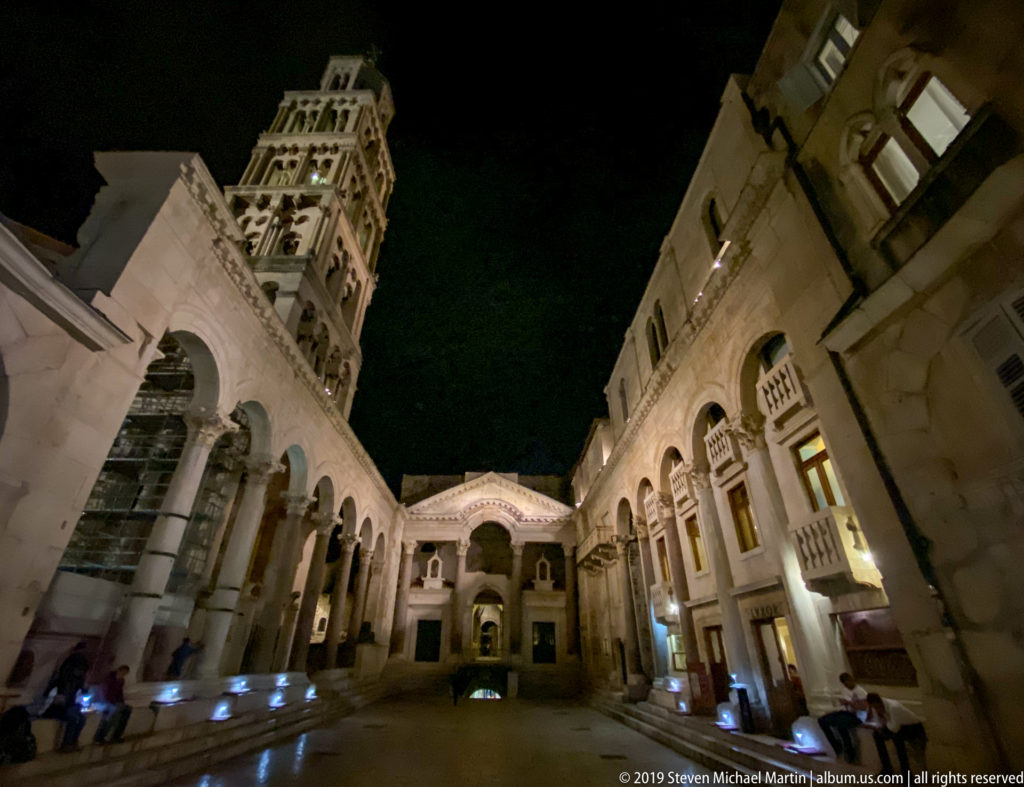 Illuminated stone arcades and buildings around the Peristyle at night, with the Cathedral of Saint Domnius bell tower rising above.