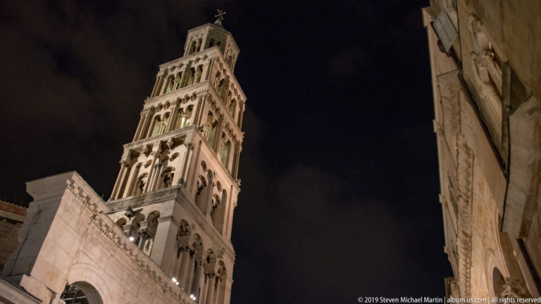 Tall illuminated Saint Domnius bell tower rises between dark stone building walls in a steep low-angle night view.