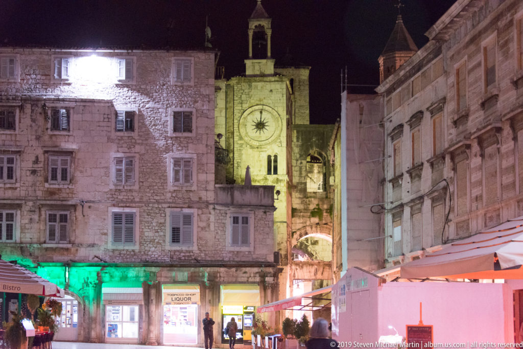 Illuminated Clock Tower and Iron Gate beyond buildings and cafés on People’s Square at night.