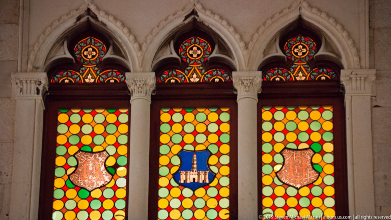 Three pointed stained-glass windows with colorful roundels and heraldic shields, separated by carved pale-stone columns.