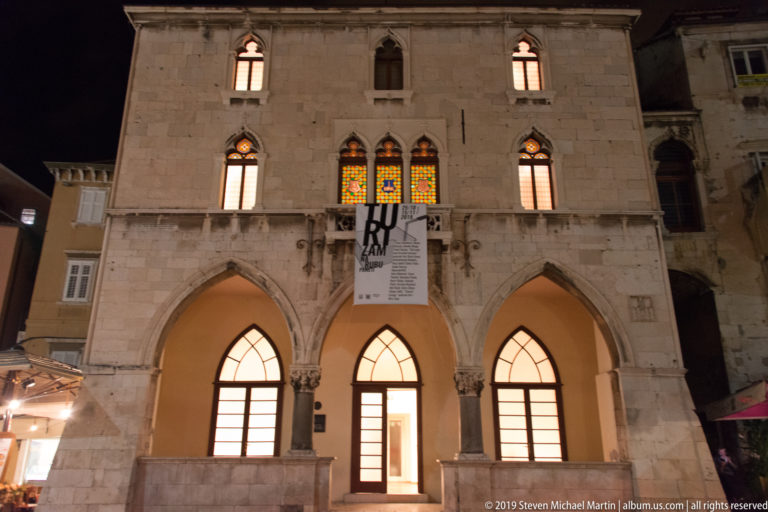 Illuminated Old Town Hall façade on People’s Square with pointed arches, traceried windows, and an exhibition banner.