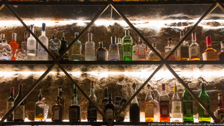 Two backlit shelves filled with liquor bottles in a black geometric frame against rough exposed stone.