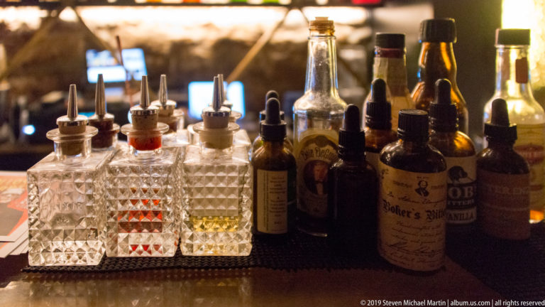 Cut-glass bottles with pour spouts, small droppers, bitters, and other cocktail ingredients arranged across a dim bar counter.