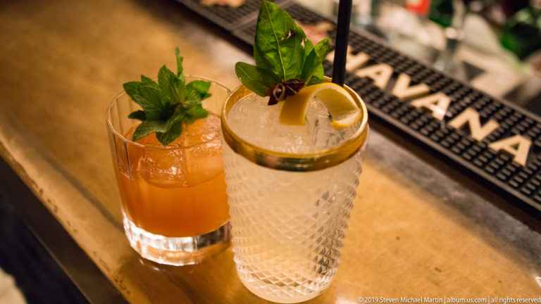 Two cocktails on a bar: an amber iced drink with mint and a pale highball garnished with citrus and spices.