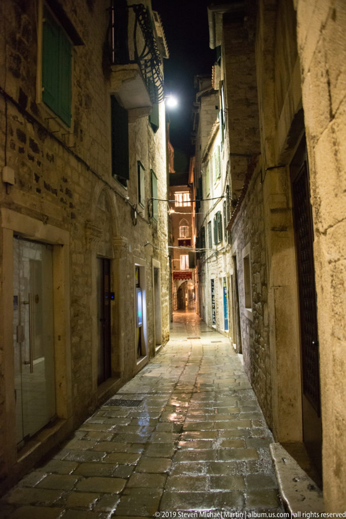 Empty narrow stone-paved alley at night, lined with rough masonry buildings, shutters, doors, and projecting upper stories.