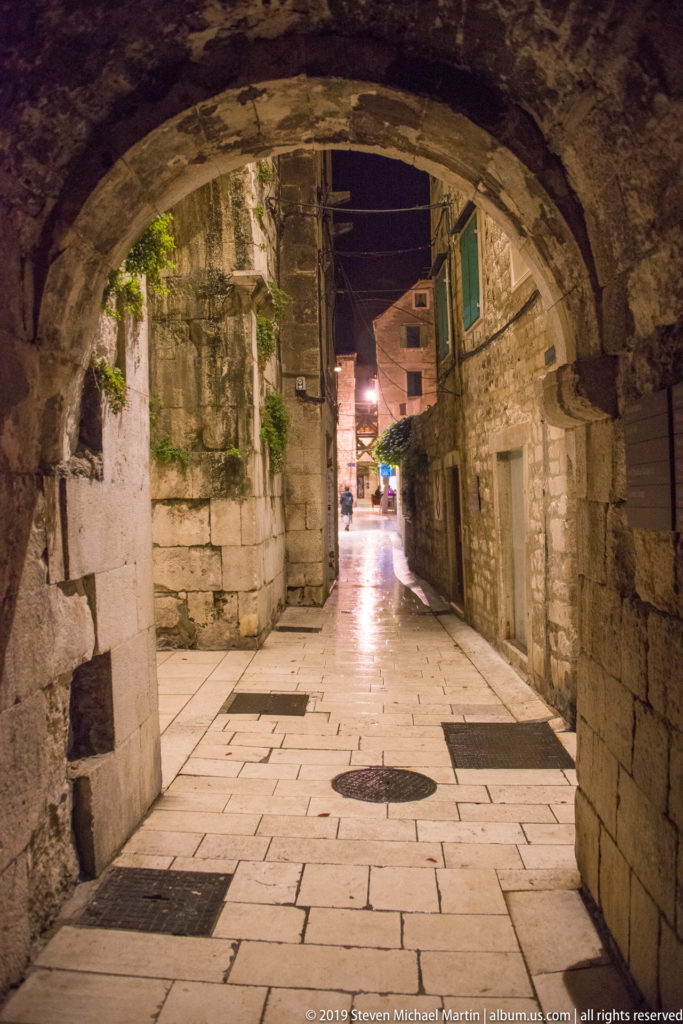 Large stone arch framing a narrow illuminated lane between old masonry buildings, with pale paving reflecting the night lights.