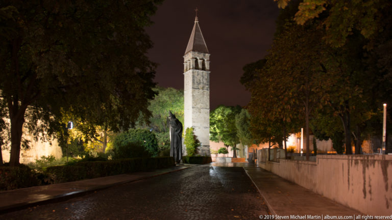 Tall narrow stone bell tower with arched openings and a pointed roof, illuminated beside a dark tree-lined walkway.