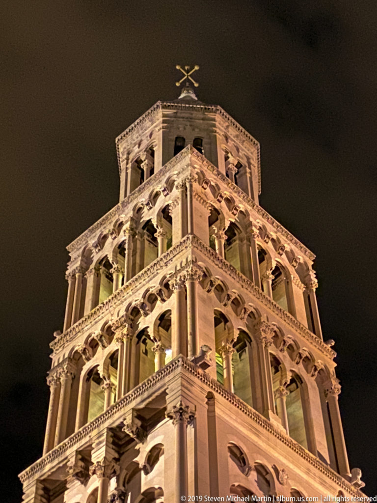 Illuminated upper tiers of Saint Domnius bell tower with stacked arcades and slender stone columns against a black night sky.