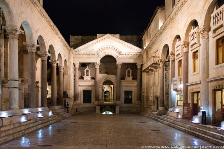 Wide paved Peristyle courtyard at night with illuminated colonnades, central arch, and historic buildings enclosing both sides.