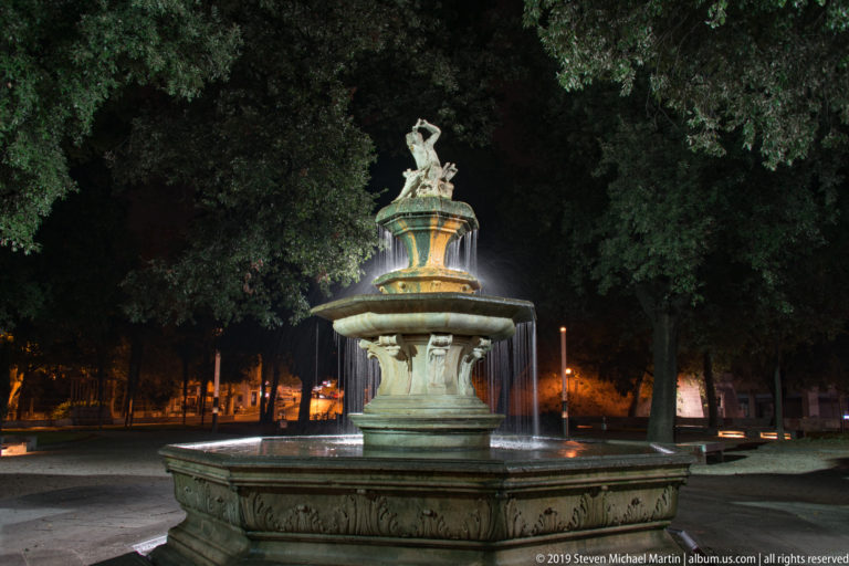 Full illuminated tiered fountain with carved lower basin and sculptural figures, surrounded by mature trees in a paved park.