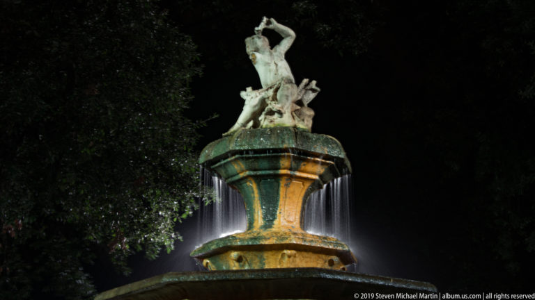 Illuminated sculptural fountain with water spilling from an upper basin and pale figures silhouetted against dark trees.