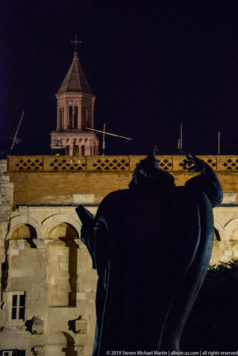 Close nighttime silhouette of the monumental Gregory of Nin statue, with raised arm and heavy robes against illuminated stone buildings.
