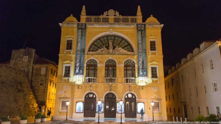 Illuminated Croatian National Theatre in Split with arched entrances, central glazed arch, balconies, and banners on its ornate façade.