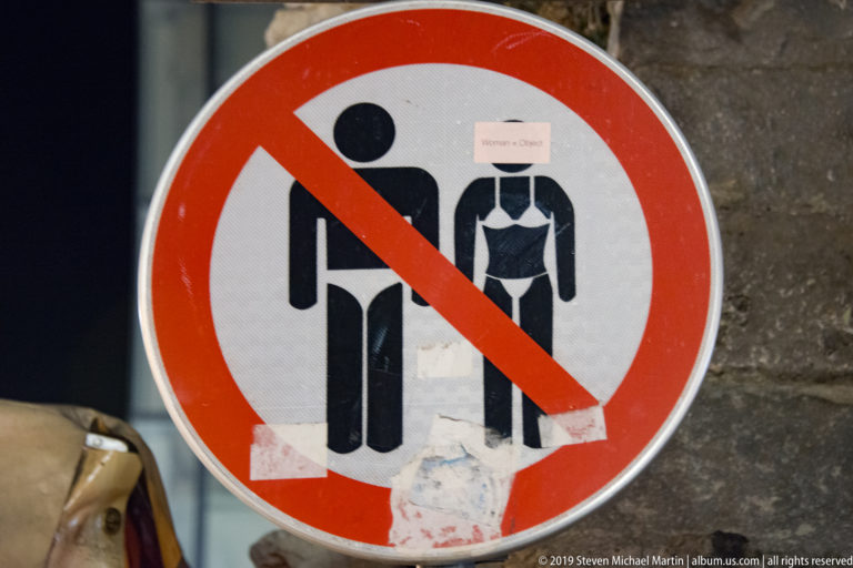 Weathered circular prohibition sign with male and female pictograms crossed by a red diagonal stripe and partly covered by stickers.