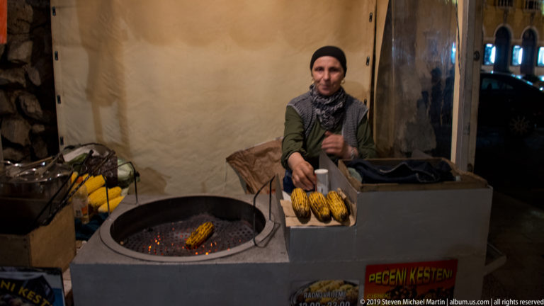 Vendor behind a charcoal brazier holding grilled corn, with a sign for roasted chestnuts on the street-food cart.