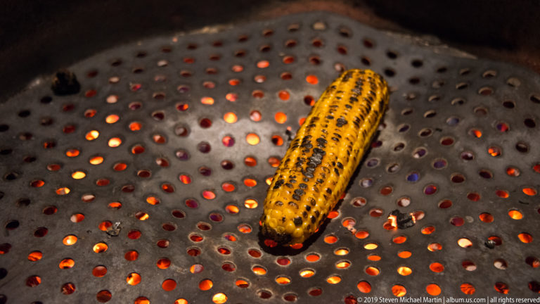 Ear of corn roasting over glowing coals on a perforated round metal brazier.