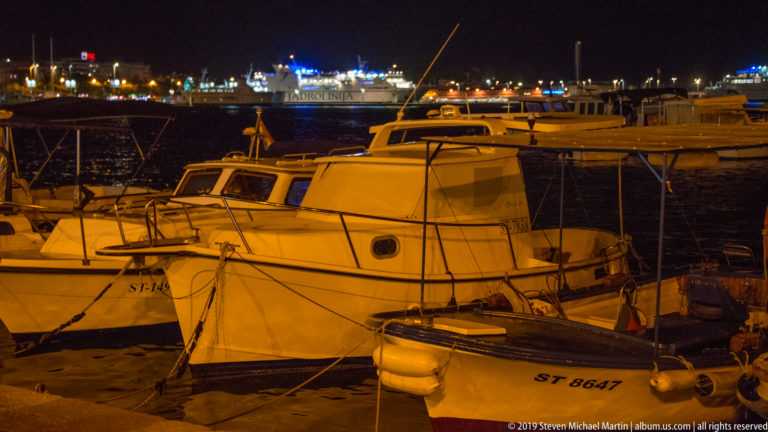 Small white cabin motorboats tied at the quay with ropes and fenders, with larger illuminated vessels across the harbor.