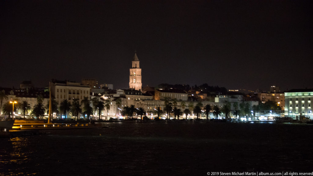 Split waterfront glowing across the harbor at night, with palm trees and the Cathedral of Saint Domnius bell tower above the skyline.