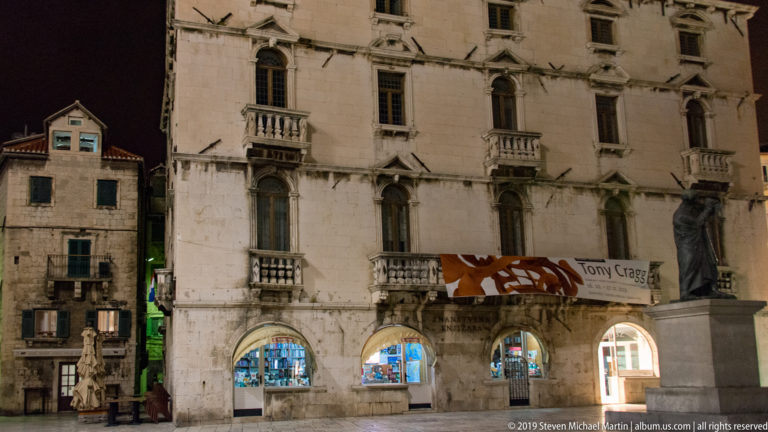 Illuminated ornate stone façade with carved windows, small balconies, and arched ground-floor shopfronts beside an older building.