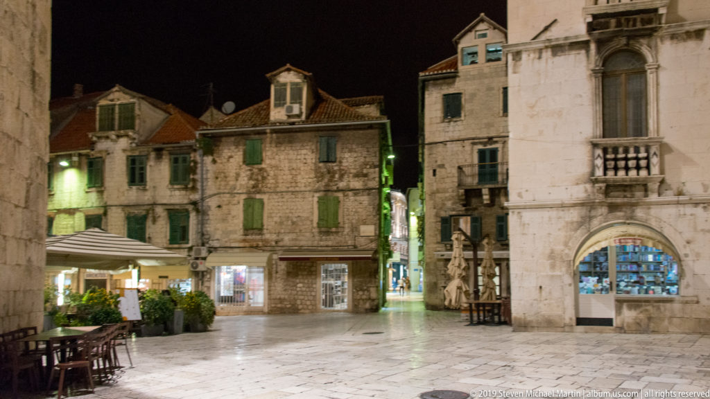 Quiet paved square at night bordered by old stone buildings, shuttered windows, shops, and a narrow illuminated passage.