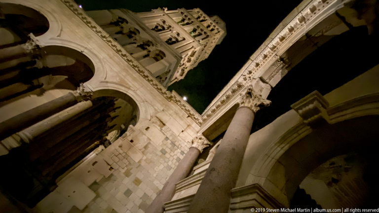 Extreme low-angle night view of Saint Domnius bell tower rising above diagonal stone columns and arches of the Peristyle.