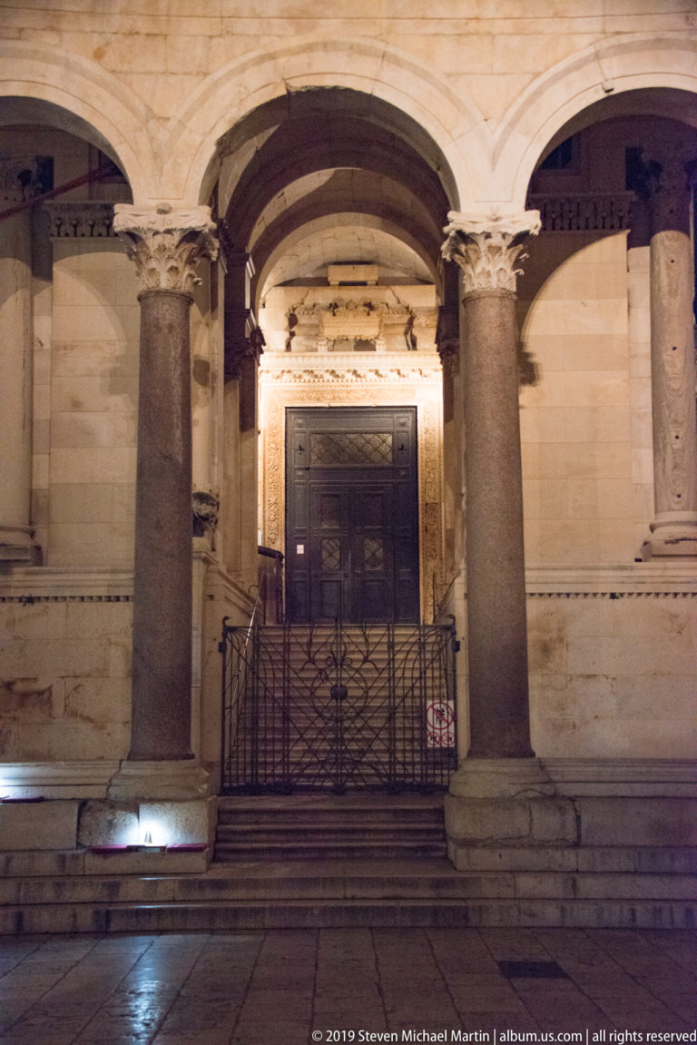 Symmetrical night view of Saint Domnius Cathedral entrance between two massive columns, with steps and a wrought-iron barrier in front.