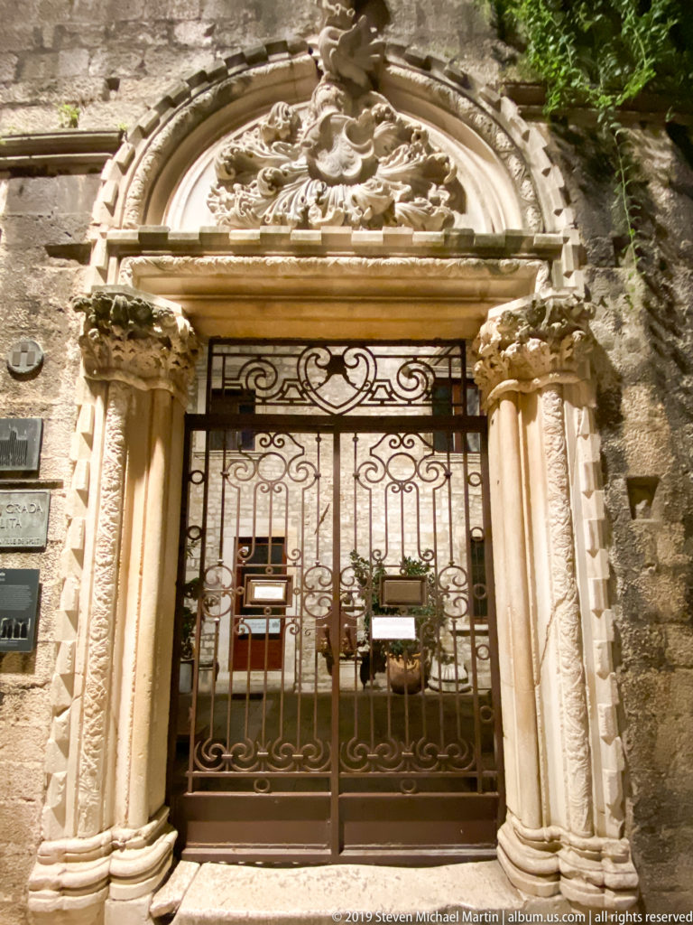 Full ornate stone portal with carved side supports, wrought-iron gate, and sculpted rounded pediment illuminated at night.
